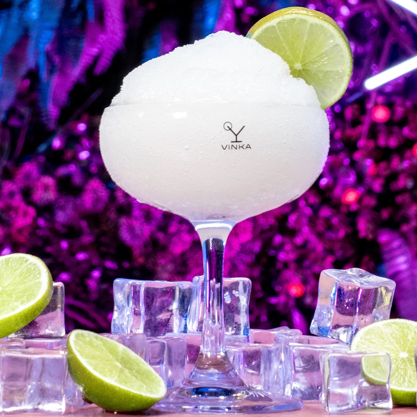 Frozen VINKA Margarita with a pinky purple background. Lime garnishes in a VINKA Stemmed glass.