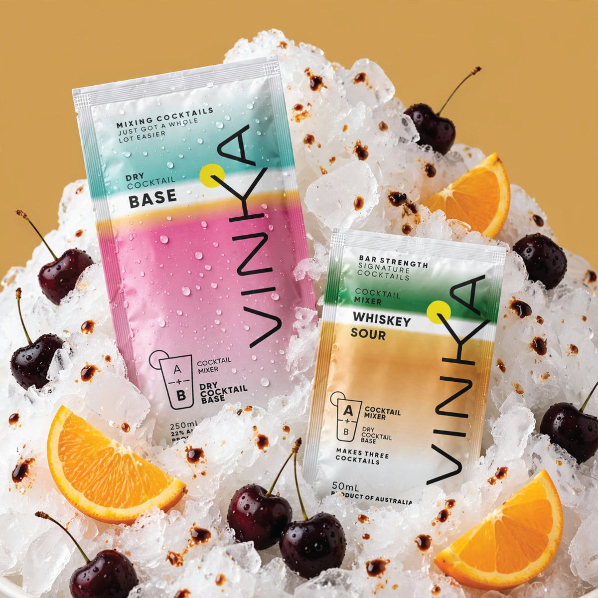 Two VINKA cocktail base packets, one labeled 'Mixino Cocktails' and the other 'Whiskey Sour', displayed on a bed of crushed ice with garnished cherries and orange slices against a warm tan background.