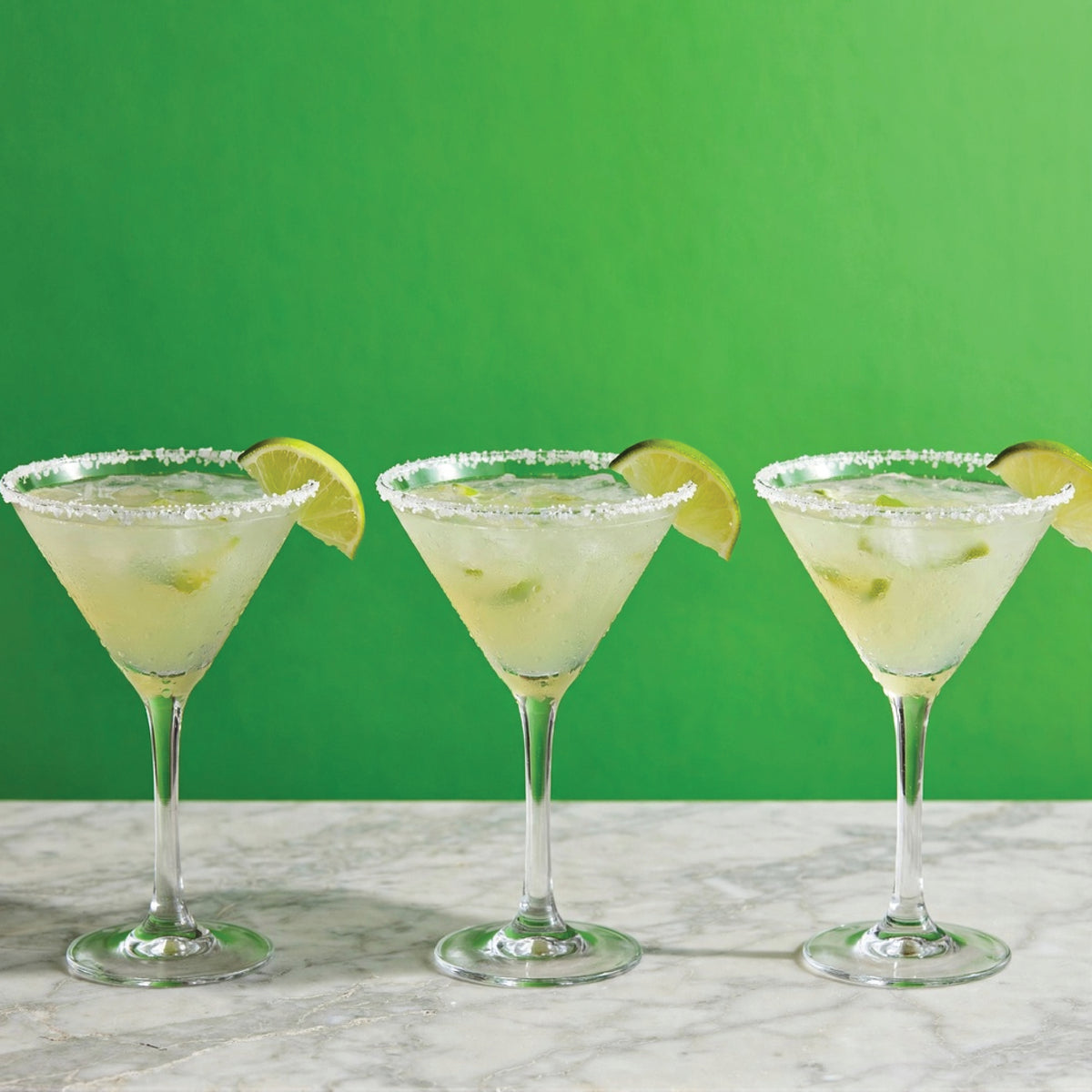 Three margarita glasses with salted rims and lime wedges, filled with a pale yellow drink, arranged on a marble surface against a solid green background.