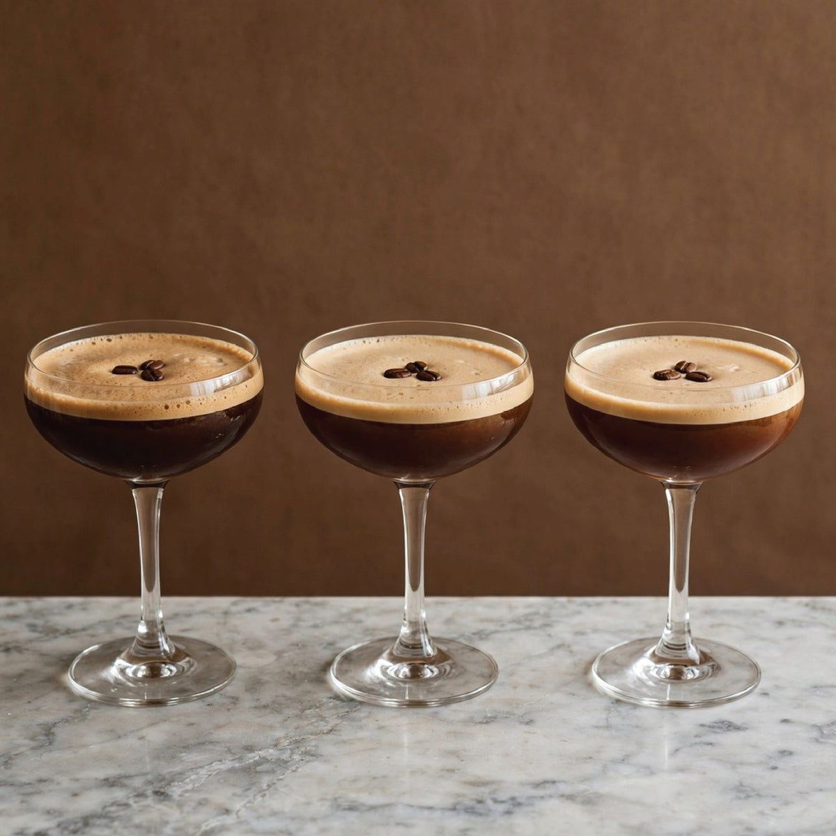 Three stemmed coupe glasses filled with an espresso-based cocktail, each topped with a thick cream layer and a small dark garnish, arranged on a marble surface against a brown background.