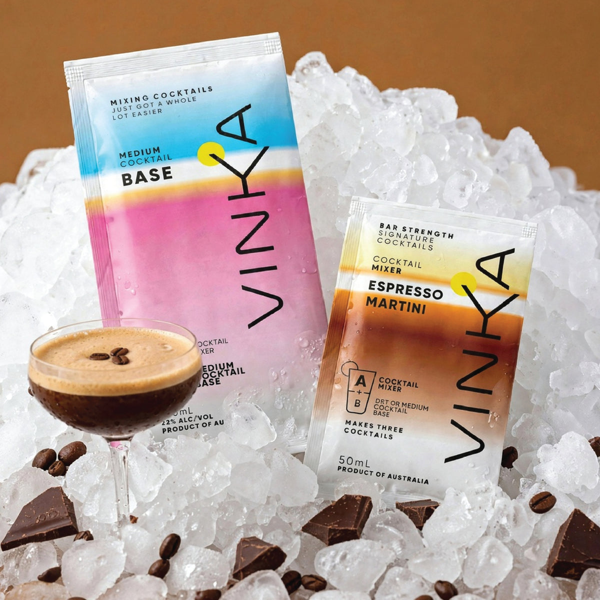 Two VINKA cocktail sachets, one labeled 'Base' and the other 'Espresso Martini', displayed among crushed ice, chocolate chunks, and a prepared cocktail glass on a warm brown background.