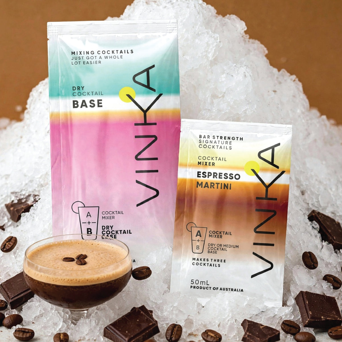 Two VINKA cocktail mix packets, one labeled 'Dry Cocktail Base' and the other 'Espresso Martini', displayed on crushed ice with chocolate pieces and a small espresso drink in the foreground against a warm brown background.