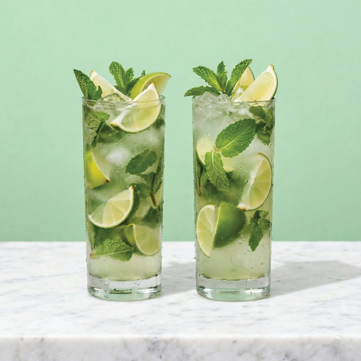 Two tall glasses of a mint and lime cocktail, filled with ice, fresh mint leaves, and lime wedges, set on a white marble surface against a light green background.