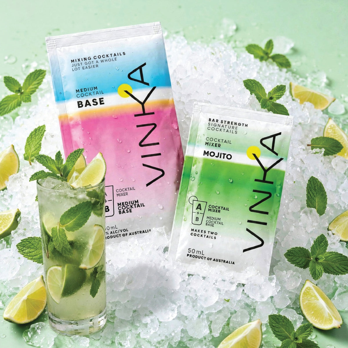 Two VINKA cocktail mix sachets, one labeled 'Base' with a pink-to-blue gradient and one labeled 'Mojito' with a green-to-white gradient, displayed on crushed ice with mint leaves and lime wedges around a glass of mixed drink.