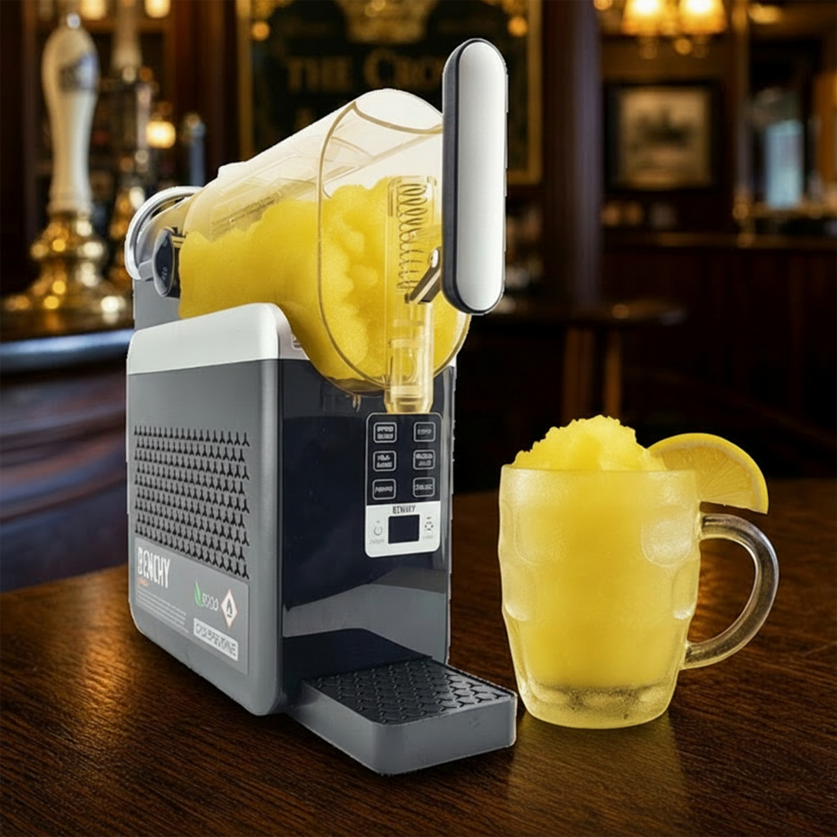 A countertop beverage machine dispensing a yellow frozen drink into a clear glass mug, with a yellow slushie and lemon garnish in the mug, set on a wooden bar counter with a blurred bar interior in the background.