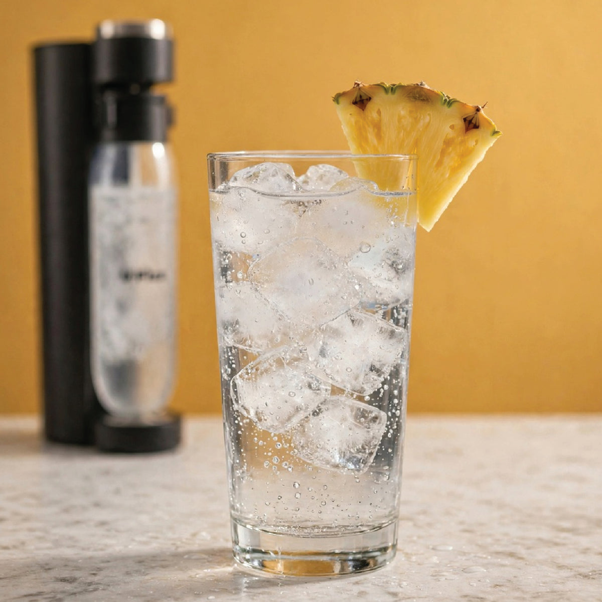 A clear glass filled with ice and carbonated liquid sits on a countertop, garnished with a pineapple slice, with a carbonated water machine blurred in the background against a warm yellow wall.