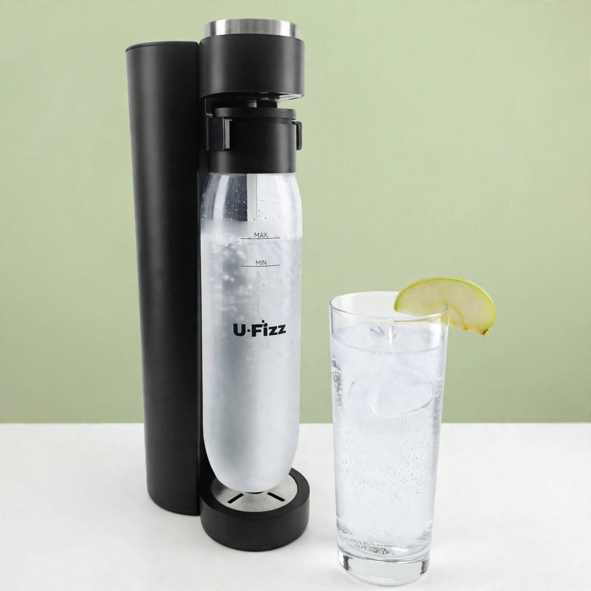 A U-Fizz carbonation device in black and clear plastic stands next to a tall glass of sparkling water with a lemon slice garnish, set against a light green background.