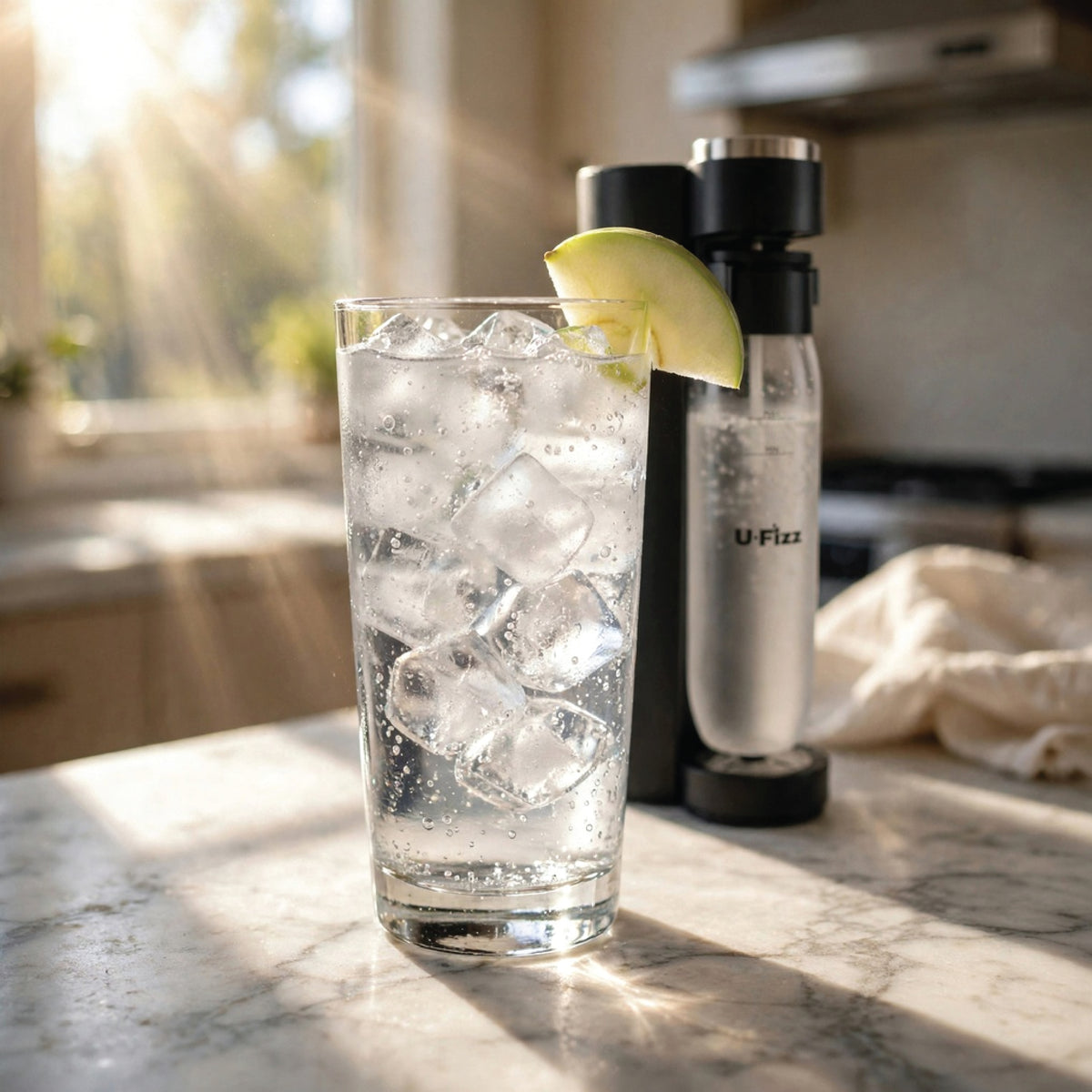 A tall clear glass filled with ice and carbonated water sits on a sunlit kitchen counter, garnished with a lime wedge, with a U-Fizz soda machine behind it.