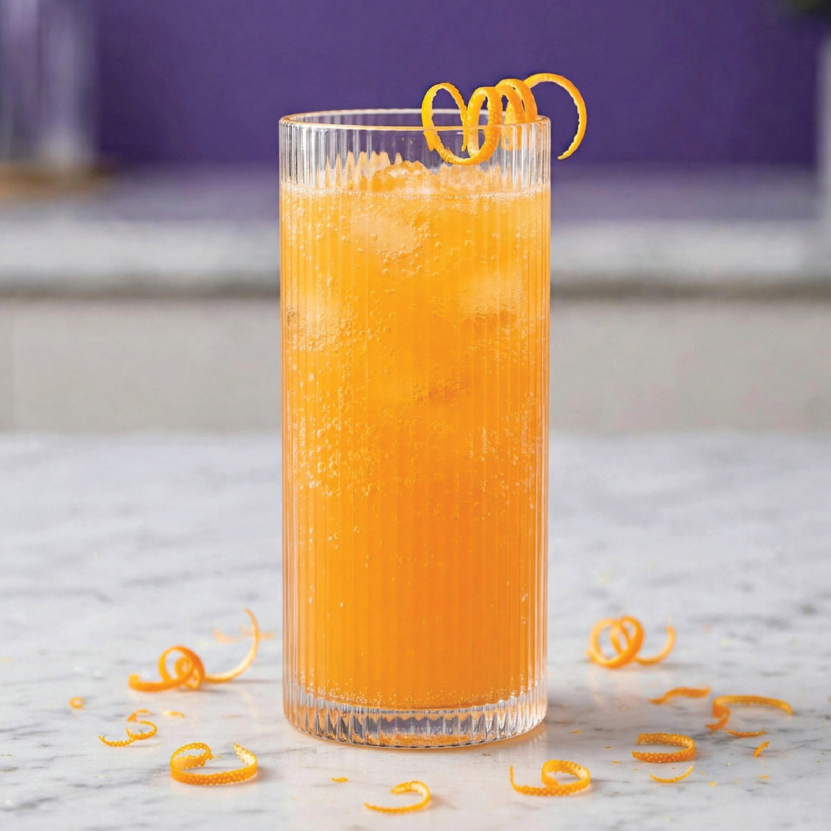 A tall ribbed clear glass filled with a bright orange beverage and ice, garnished with an orange peel curl, standing on a light marble surface with scattered orange zest curls.
