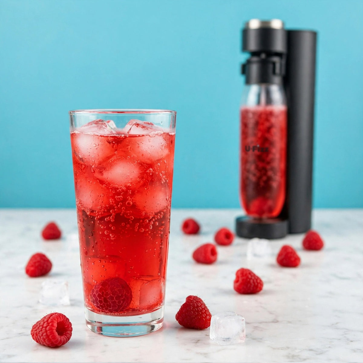 A clear glass filled with a bright red iced beverage and raspberries sits on a marble surface, with a black carbonator bottle and more raspberries in the background against a turquoise backdrop.