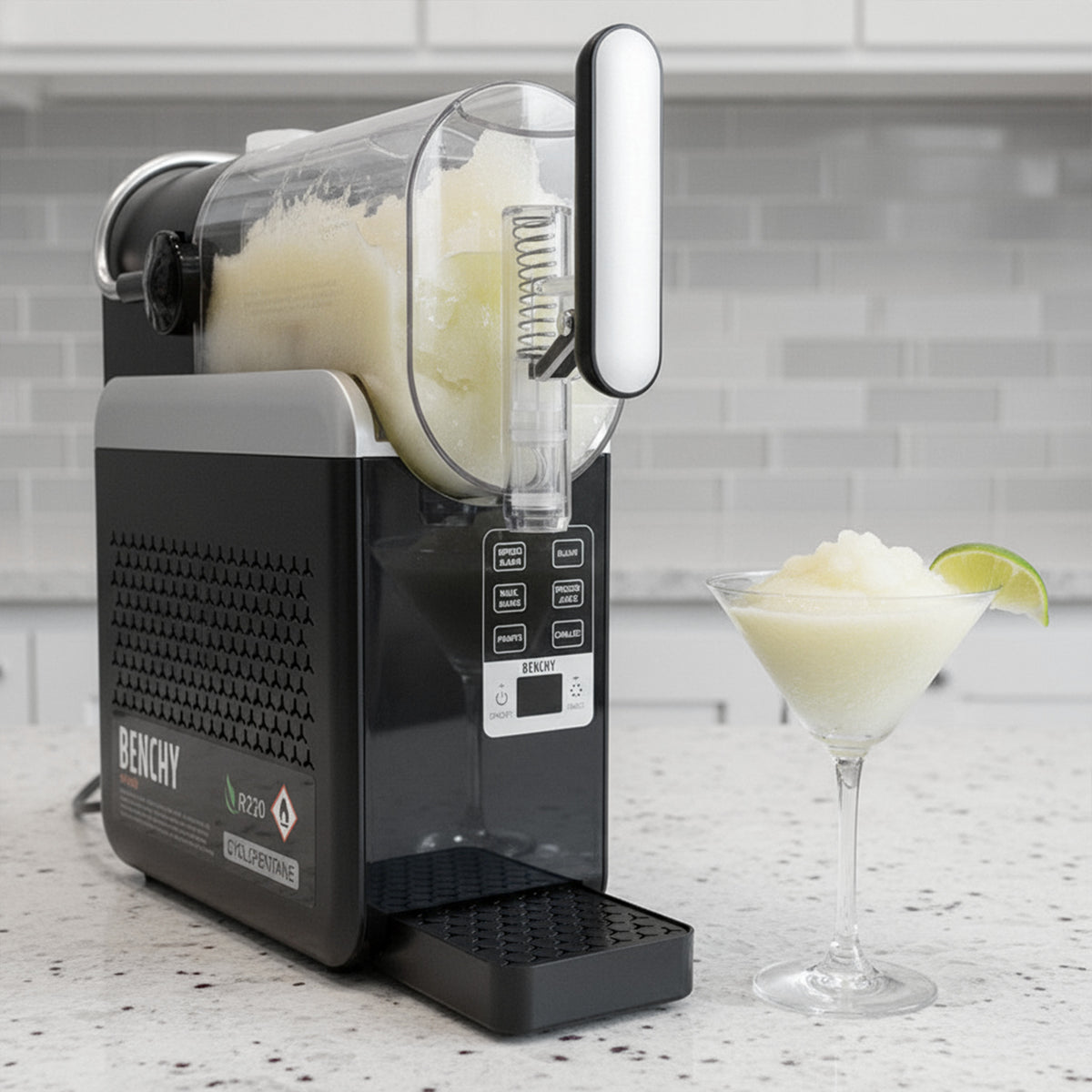 A Benchy Slushy Machine sits on a kitchen counter with a glass of Frozen Skinny Margarita VINKA Slushy Cocktail Kit (2L) and a lime wedge beside it. The background shows a light gray subway tile wall.