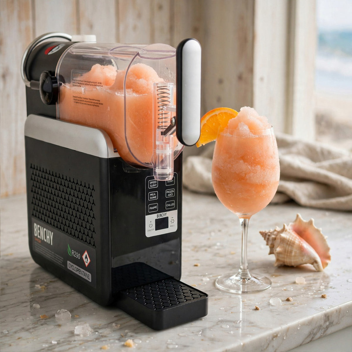 A Benchy slushie maker in black and silver, shown dispensing pink slushie into a stemmed glass garnished with an orange slice, on a marble countertop with seashells and a beach background.