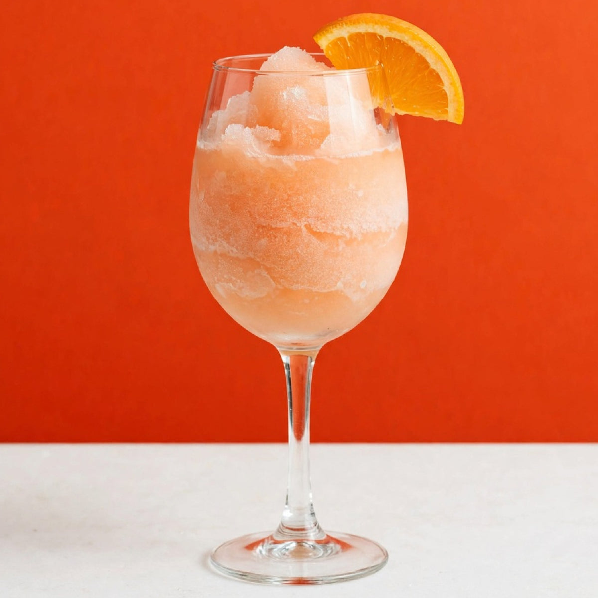 A stemmed wine glass filled with a frosted peach-orange slushie or frozen cocktail, topped with crushed ice and garnished with an orange slice, set against a solid orange background and white surface.
