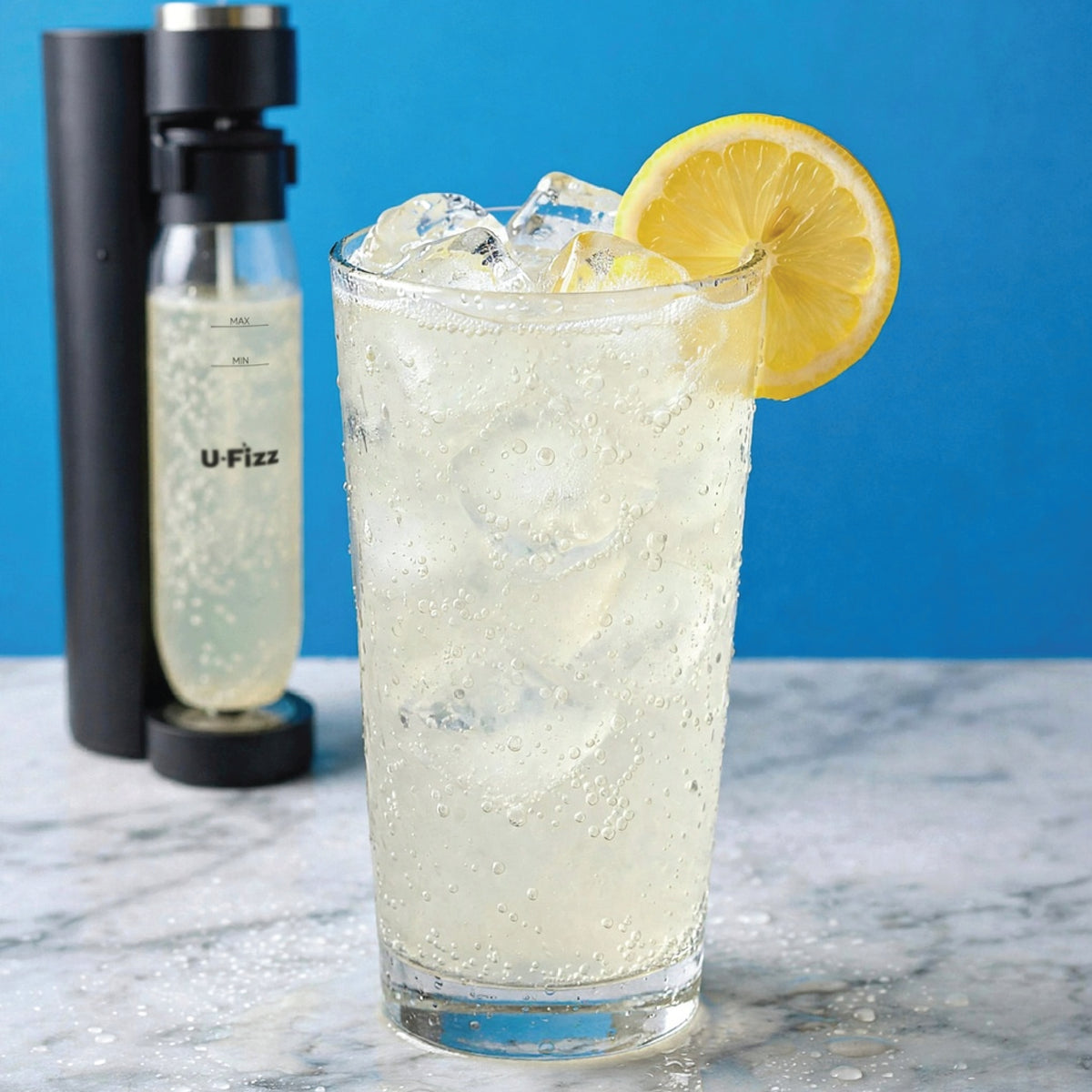 A tall clear glass filled with a bubbly pale yellow drink and ice, garnished with a lemon slice, set on a marble surface against a bright blue background. A black U-Fizz carbonator bottle stands behind it.