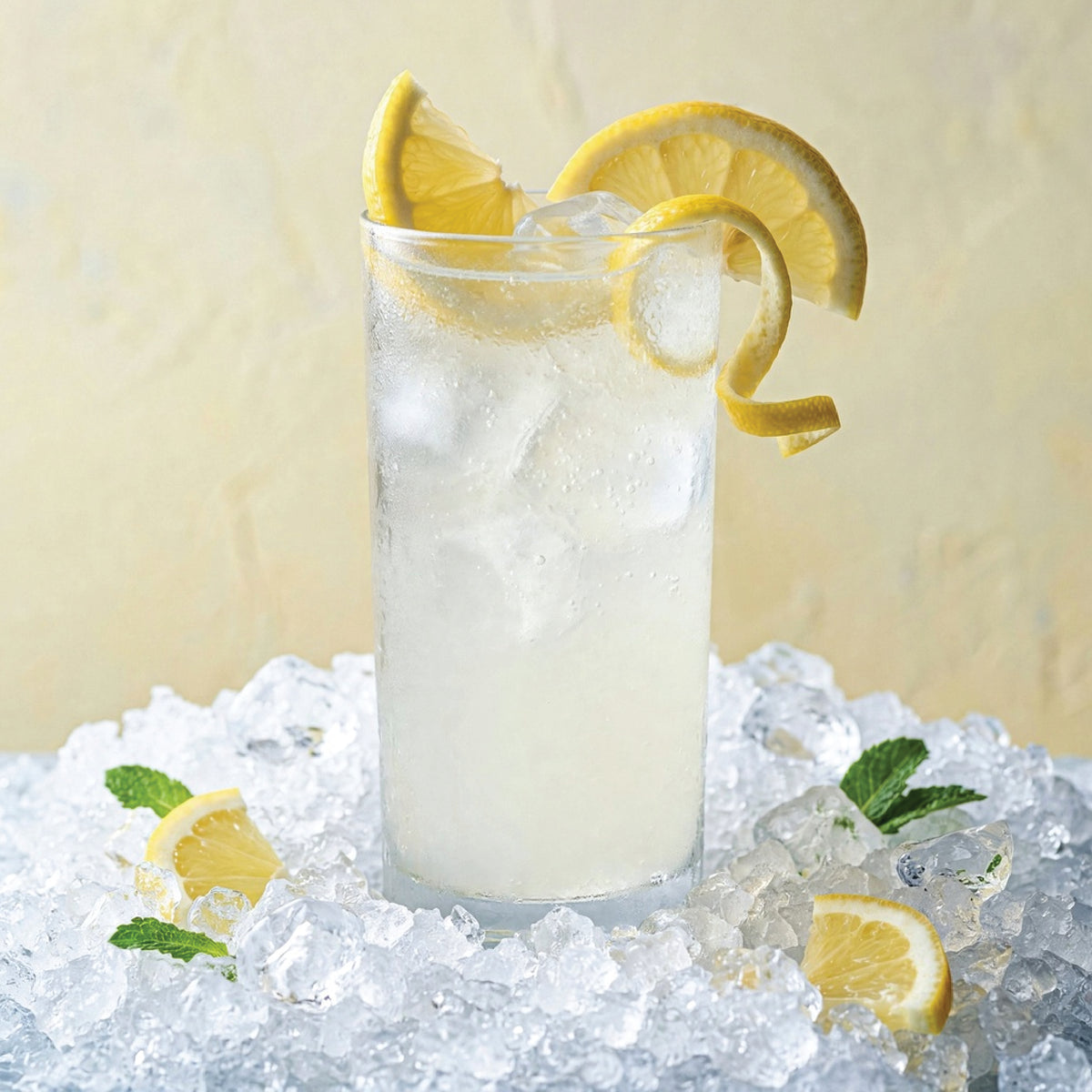A tall clear glass filled with a pale yellow iced beverage and ice cubes, garnished with lemon slices and a curled lemon peel, set in crushed ice with mint leaves against a soft beige background.