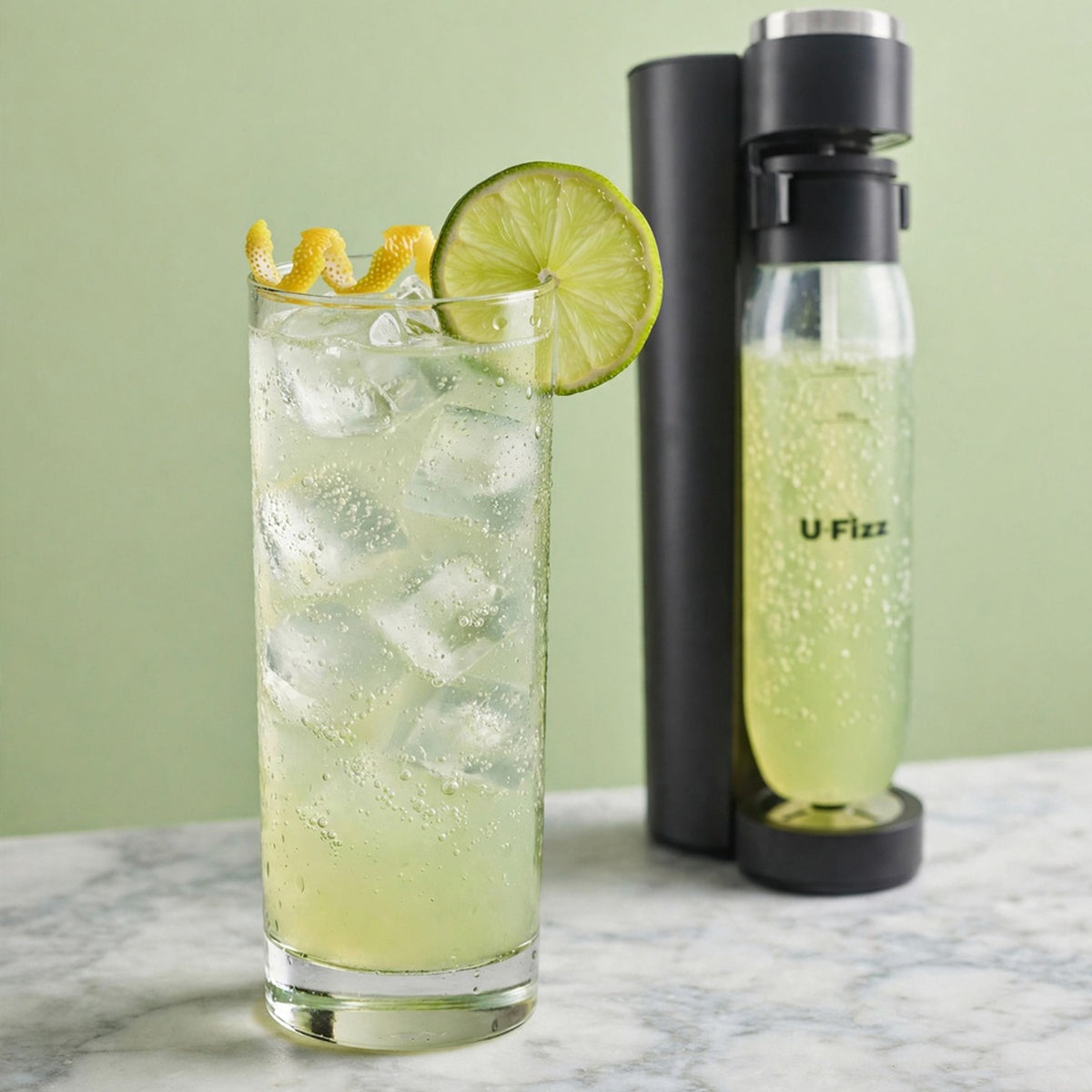 A tall glass of iced citrus drink with ice cubes, a lime wheel, and a curled lime peel garnish, shown beside a carbonation bottle and matching bottle on a marble surface against a pale green background.